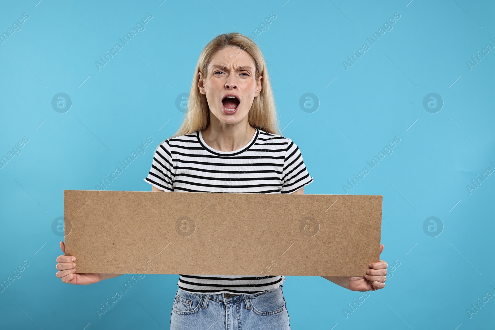 Photo of Angry woman holding blank cardboard banner on light blue background, space for text