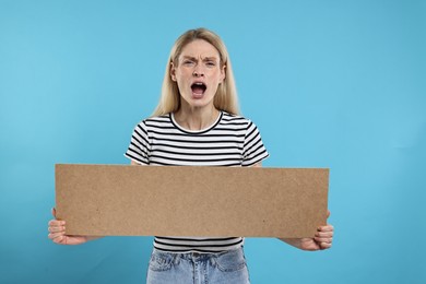 Angry woman holding blank cardboard banner on light blue background, space for text