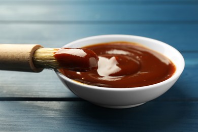 Tasty barbeque sauce in bowl and brush on blue wooden table, closeup