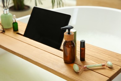 Photo of Wooden bath tray with tablet, face roller and cosmetic products on tub indoors