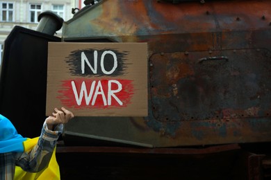 Photo of Woman wrapped in Ukrainian flag holding poster with words No War near broken military equipment on street, closeup