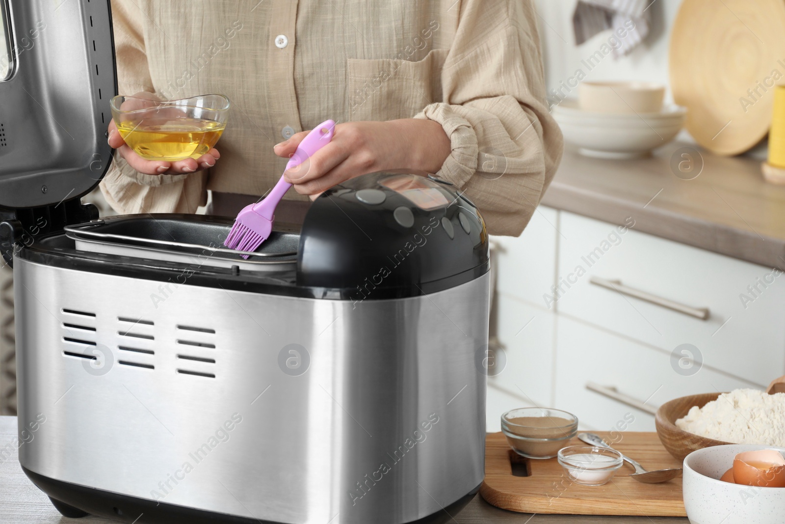 Photo of Woman spreading breadmaker pan with oil at table in kitchen, closeup