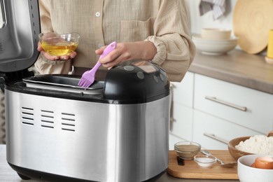 Woman spreading breadmaker pan with oil at table in kitchen, closeup