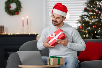 Celebrating Christmas online with exchanged by mail presents. Man in Santa hat with gift box during video call on laptop at home