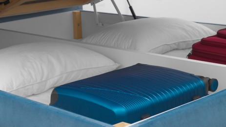 Photo of Storage drawer under bed with blue suitcase and white pillows indoors, closeup