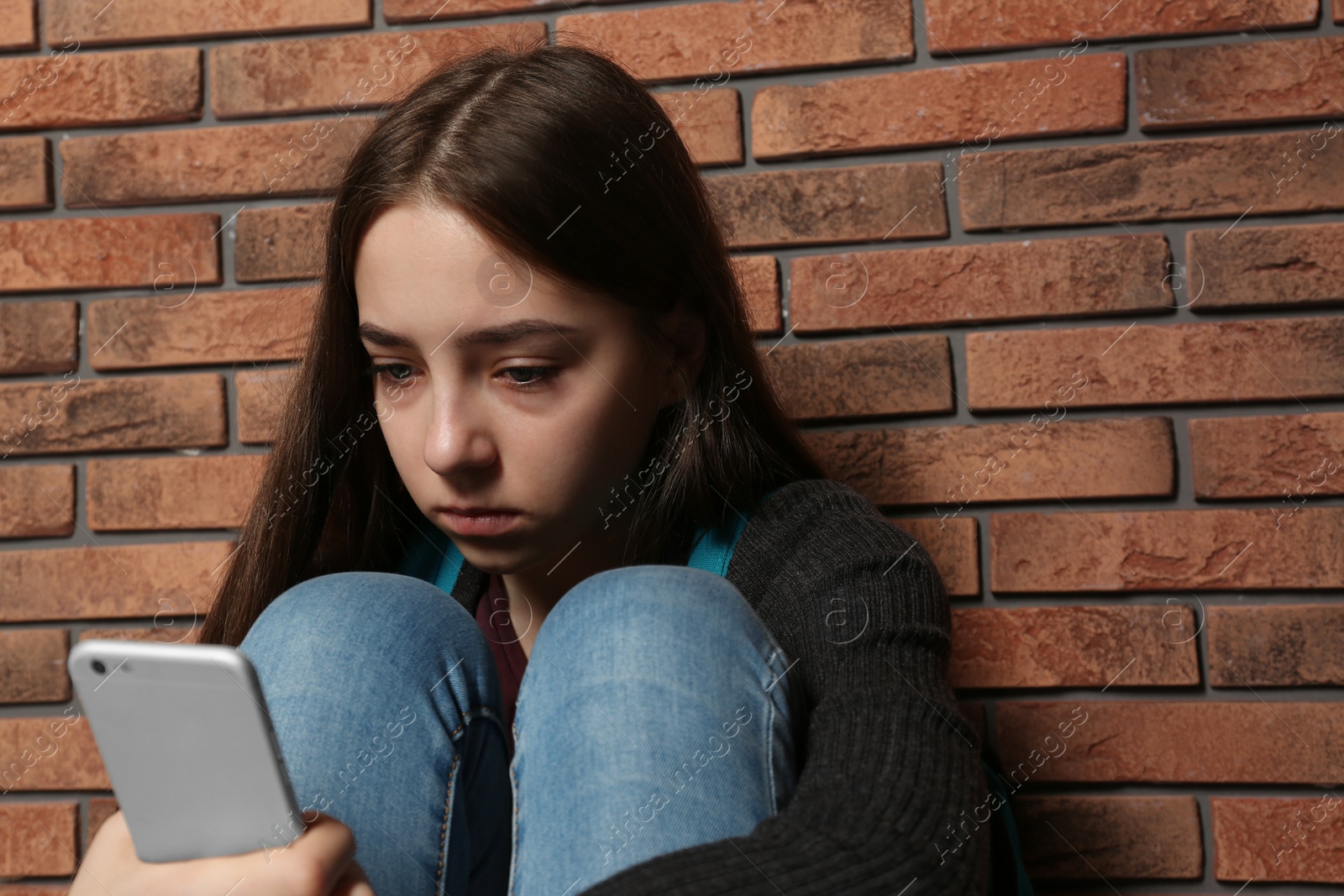 Photo of Upset teenage girl with smartphone sitting at wall indoors. Space for text