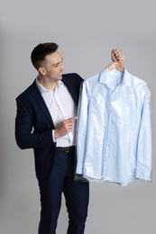 Photo of Man holding hanger with shirt in plastic bag on light grey background. Dry-cleaning service