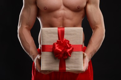 Attractive young man with muscular body holding Christmas gift box on black background, closeup