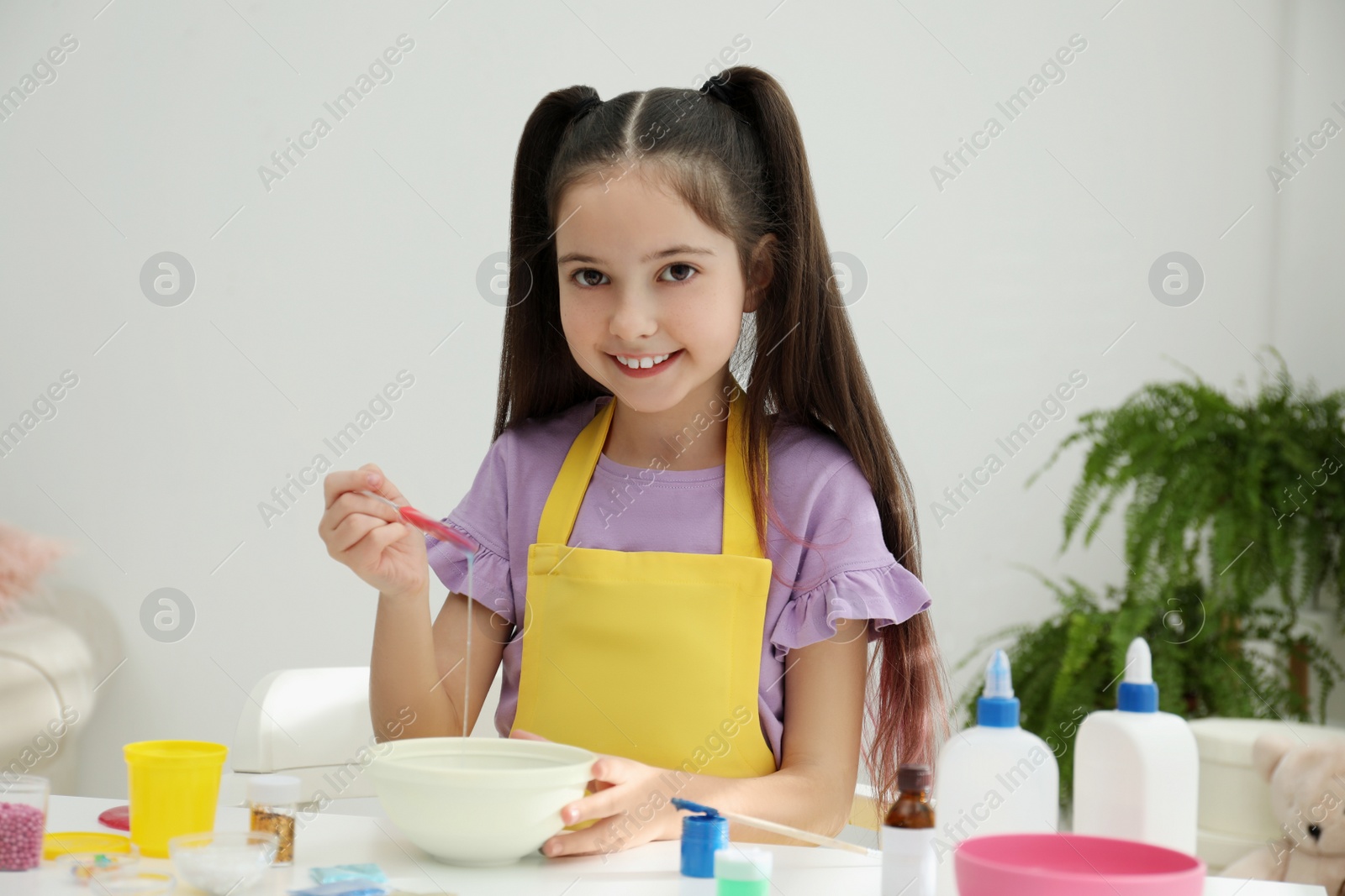 Photo of Cute little girl mixing ingredients with silicone spatula at table in room. DIY slime toy