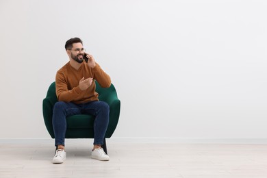 Photo of Handsome man talking on smartphone while sitting in armchair near white wall indoors, space for text