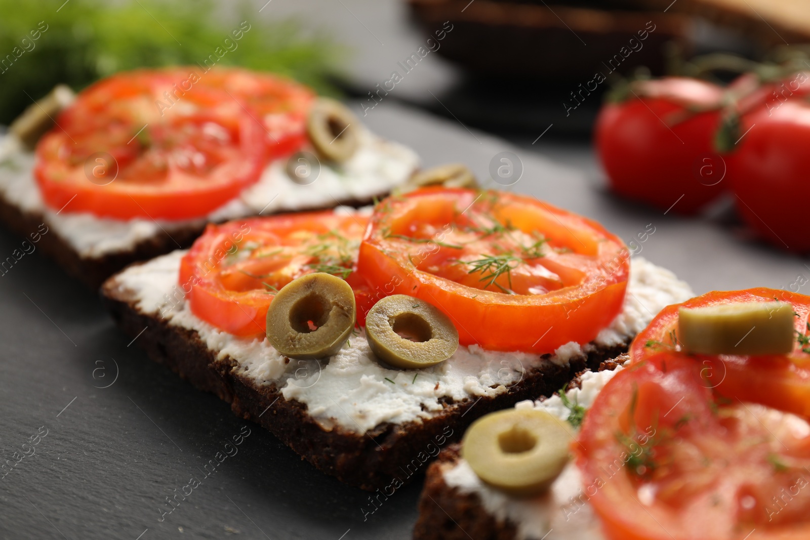 Photo of Delicious ricotta bruschettas with sliced tomatoes, olives and greens on black table, closeup