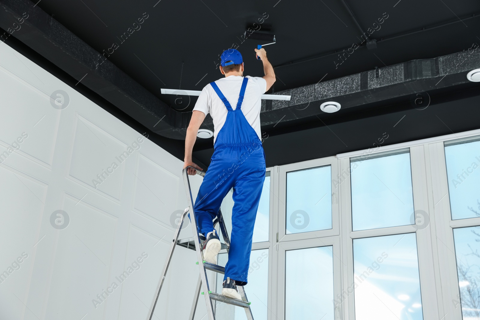 Photo of Worker in uniform painting ceiling with roller indoors, back view