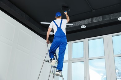 Photo of Worker in uniform painting ceiling with roller indoors, back view