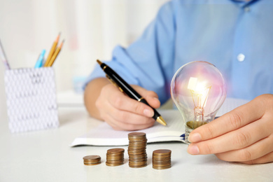 Photo of Woman with light bulb, notebook and coins at white table, closeup. Power saving