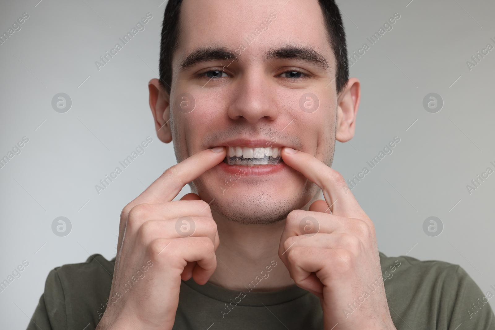 Photo of Young man applying whitening strip on his teeth against light grey background