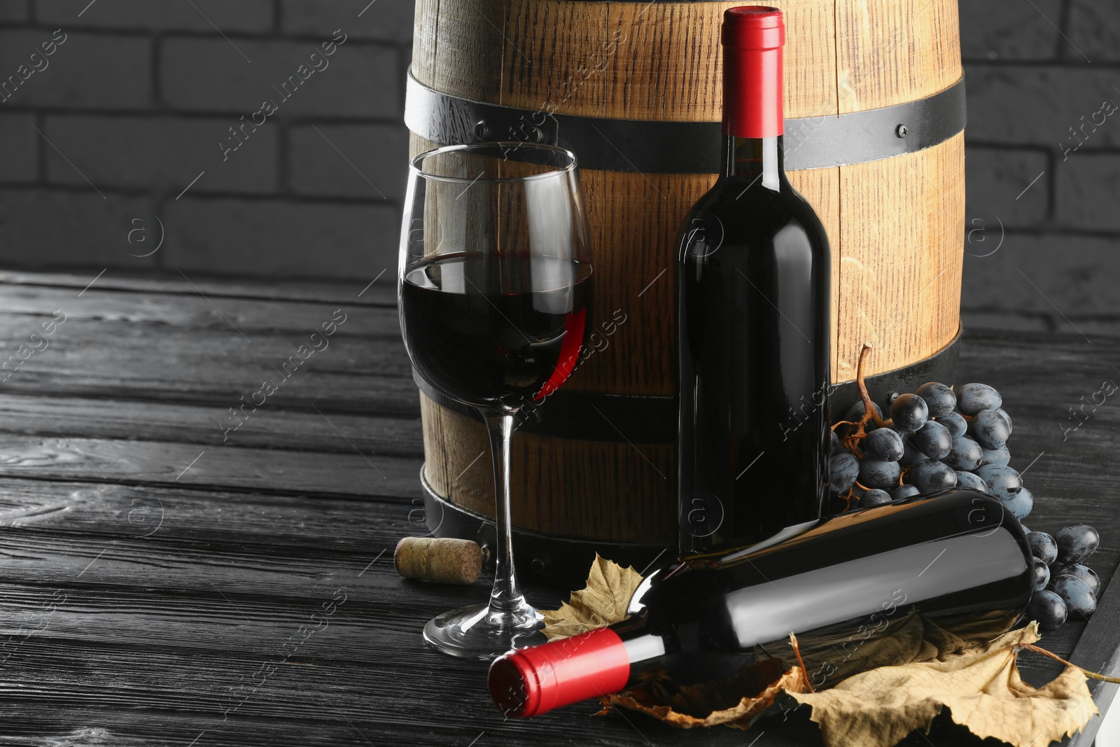 Photo of Delicious wine, wooden barrel, dry leaves and fresh grapes on black table, space for text