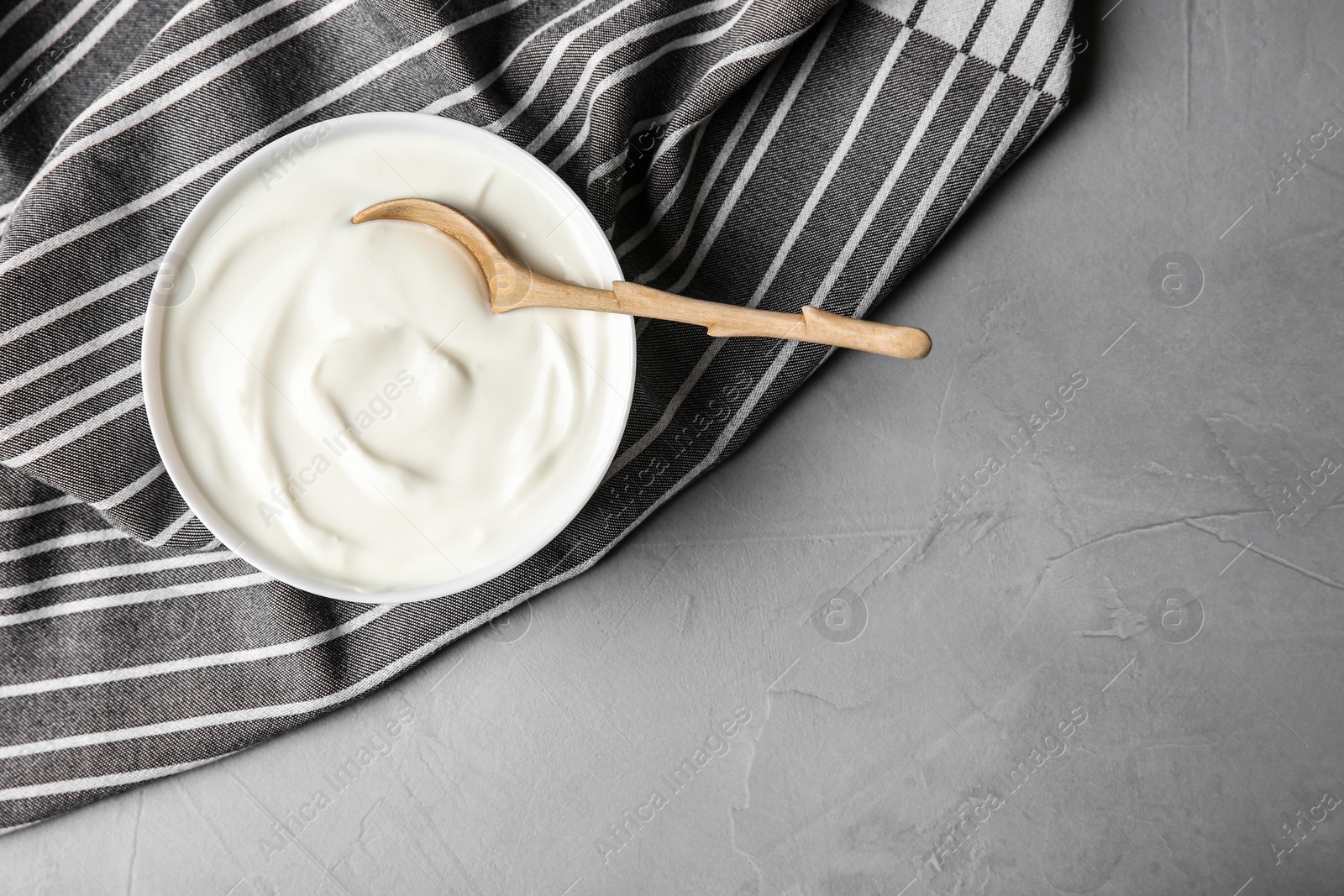 Photo of Bowl of sour cream with wooden spoon and napkin on grey table, flat lay. Space for text