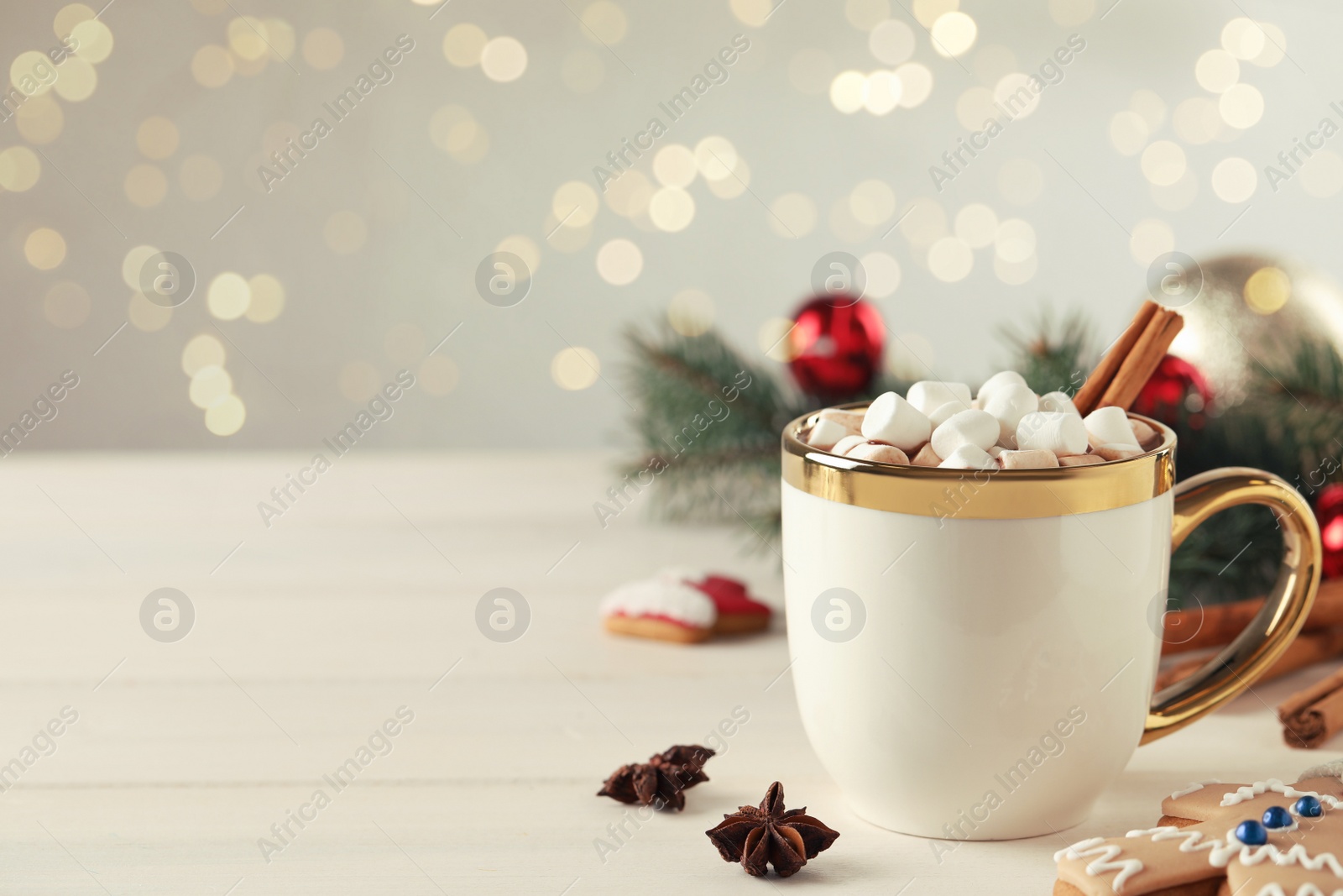 Photo of Delicious hot chocolate with marshmallows and cinnamon on white wooden table against blurred lights, space for text
