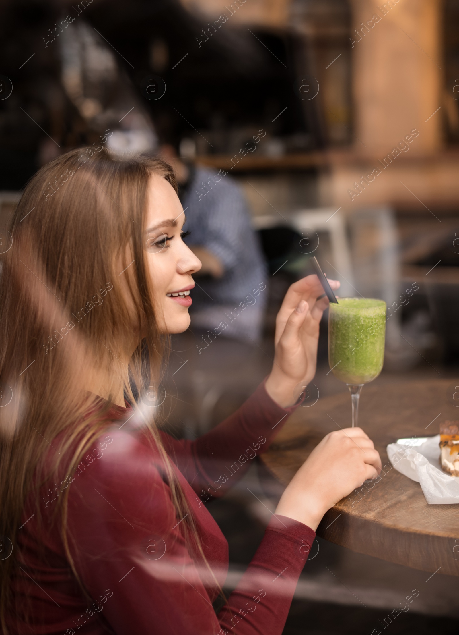 Photo of Pretty young woman with cocktail and cake at table in cafe, view from outdoors through window