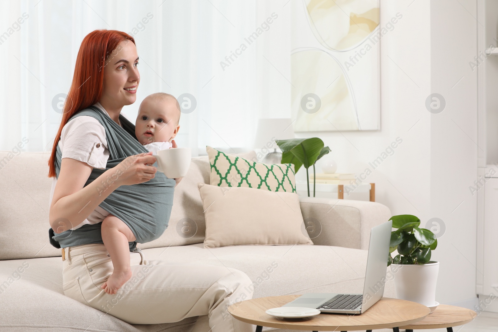 Photo of Mother with cup of drink holding her child in sling (baby carrier) at home