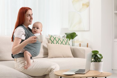 Photo of Mother with cup of drink holding her child in sling (baby carrier) at home