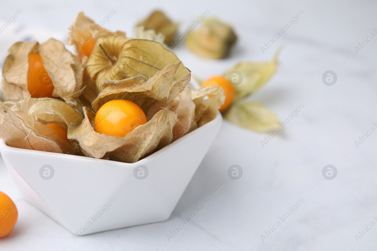 Photo of Ripe physalis fruits with calyxes in bowl on white marble table, closeup. Space for text