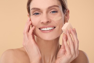 Woman blending foundation on face with makeup sponge against beige background