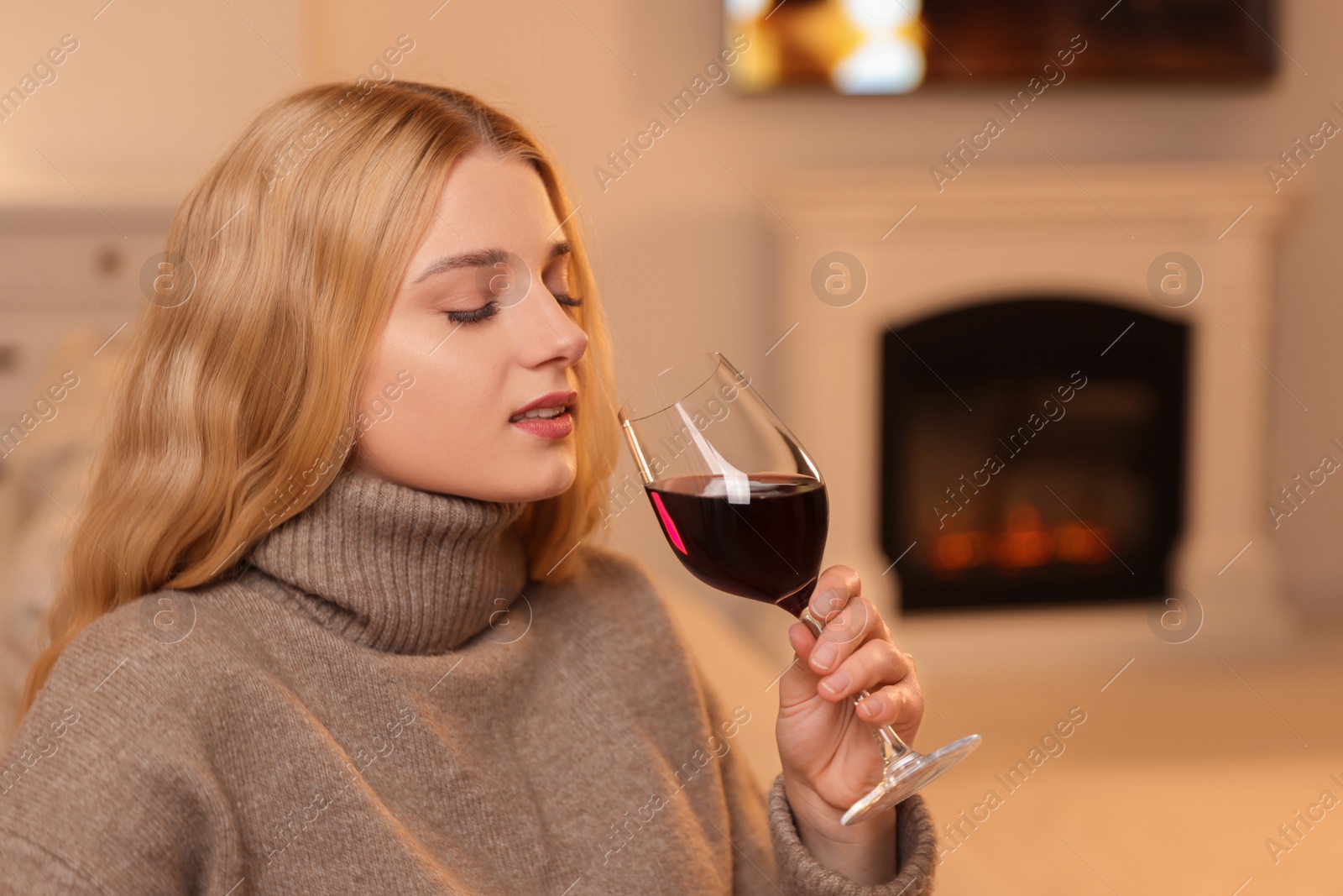 Photo of Beautiful young woman with glass of wine resting near fireplace at home