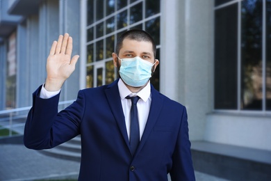 Photo of Man in protective face mask showing hello gesture outdoors. Keeping social distance during coronavirus pandemic