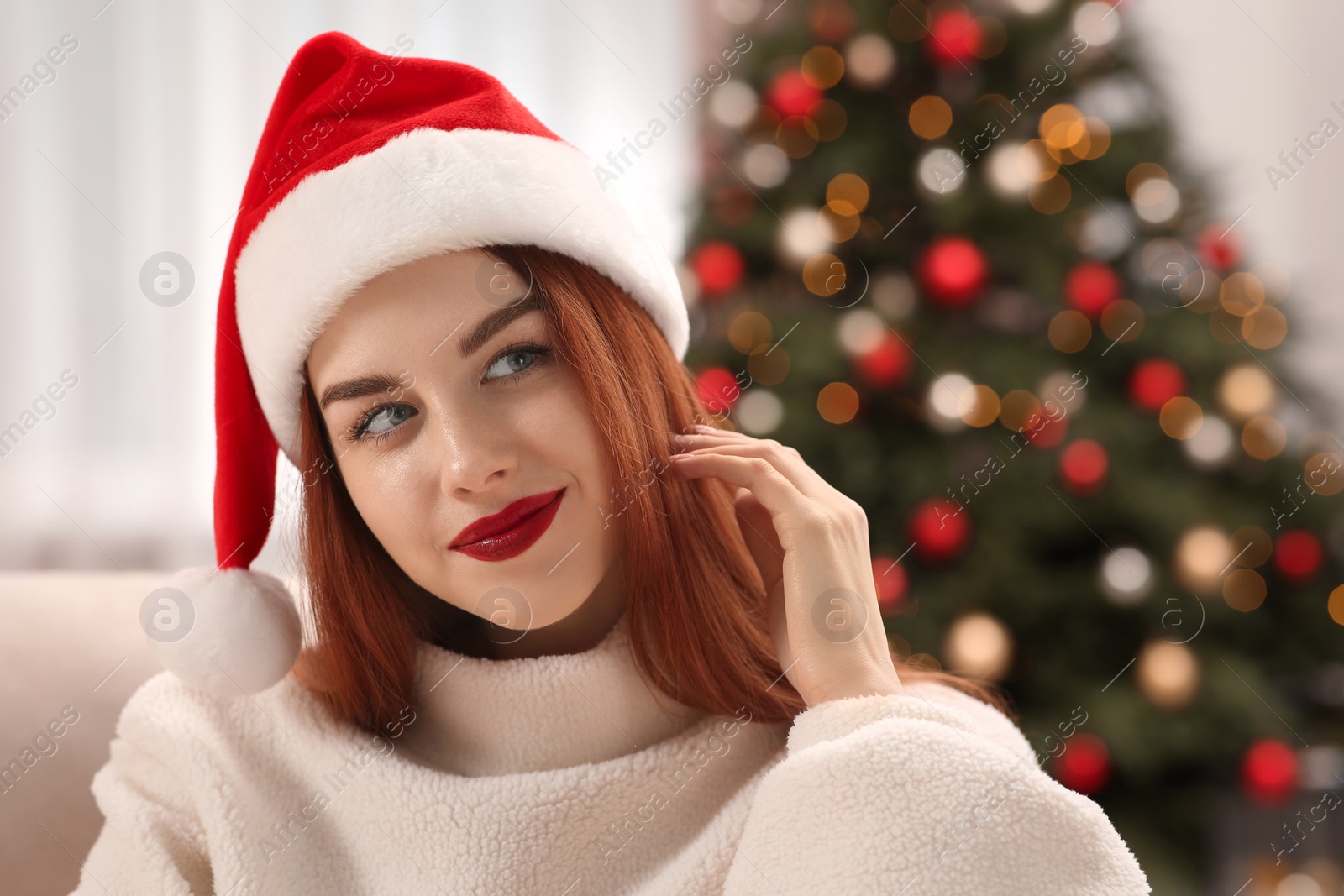 Photo of Beautiful young woman in Santa hat near Christmas tree at home, space for text