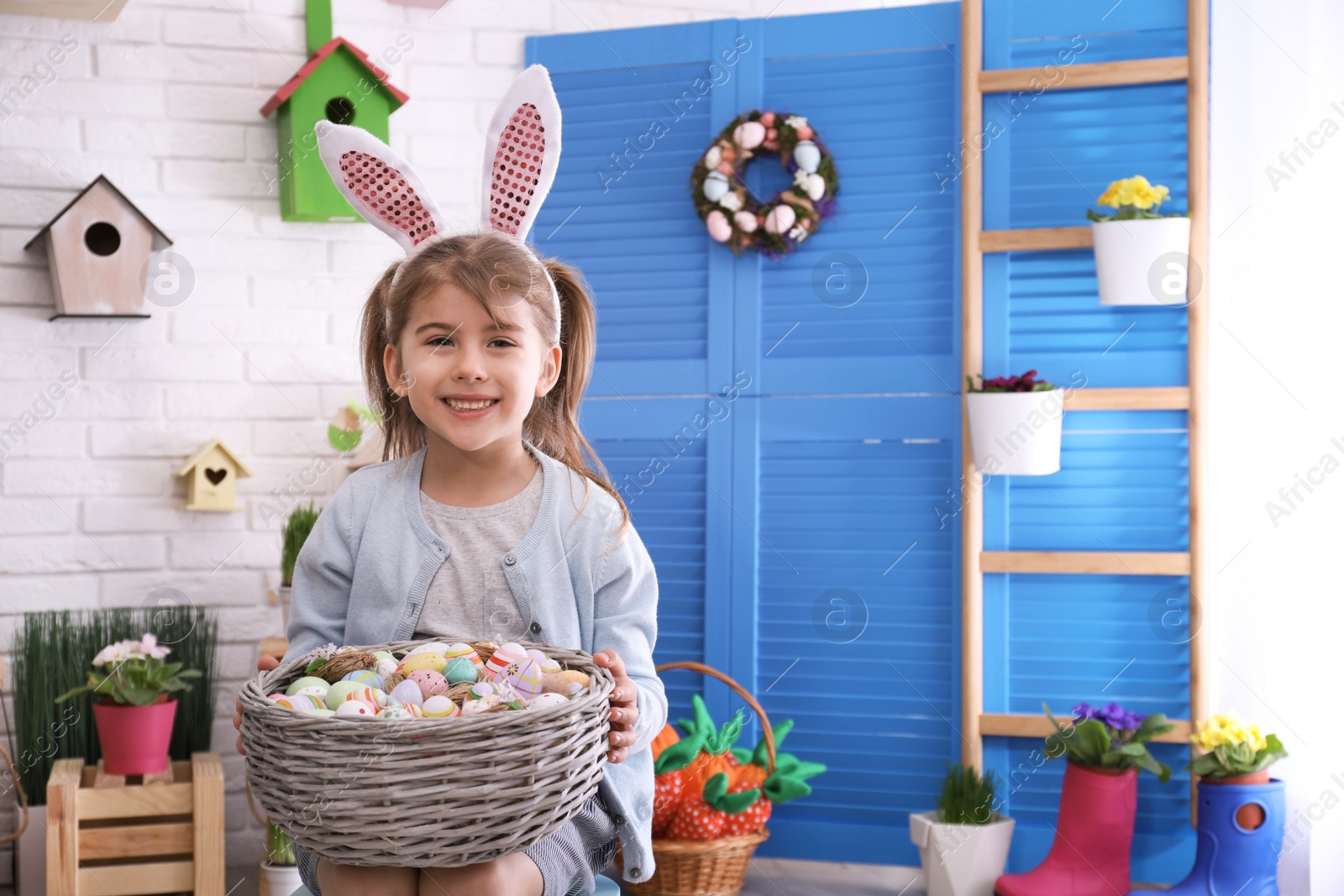 Photo of Adorable little girl with bunny ears and basket full of dyed eggs in Easter photo zone