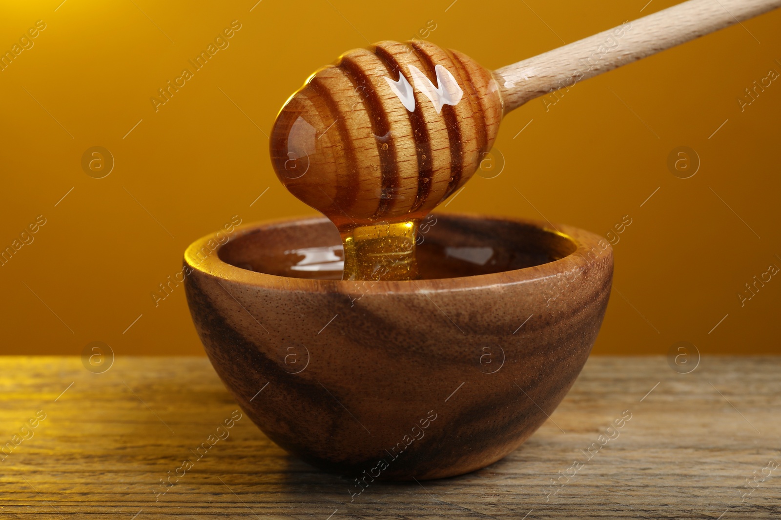 Photo of Pouring honey from dipper into bowl at wooden table against golden background, closeup