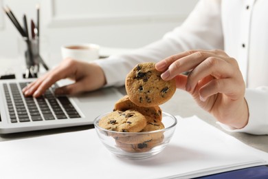 Photo of Woman taking chocolate chip cookie from bowl while working with laptop in office, closeup