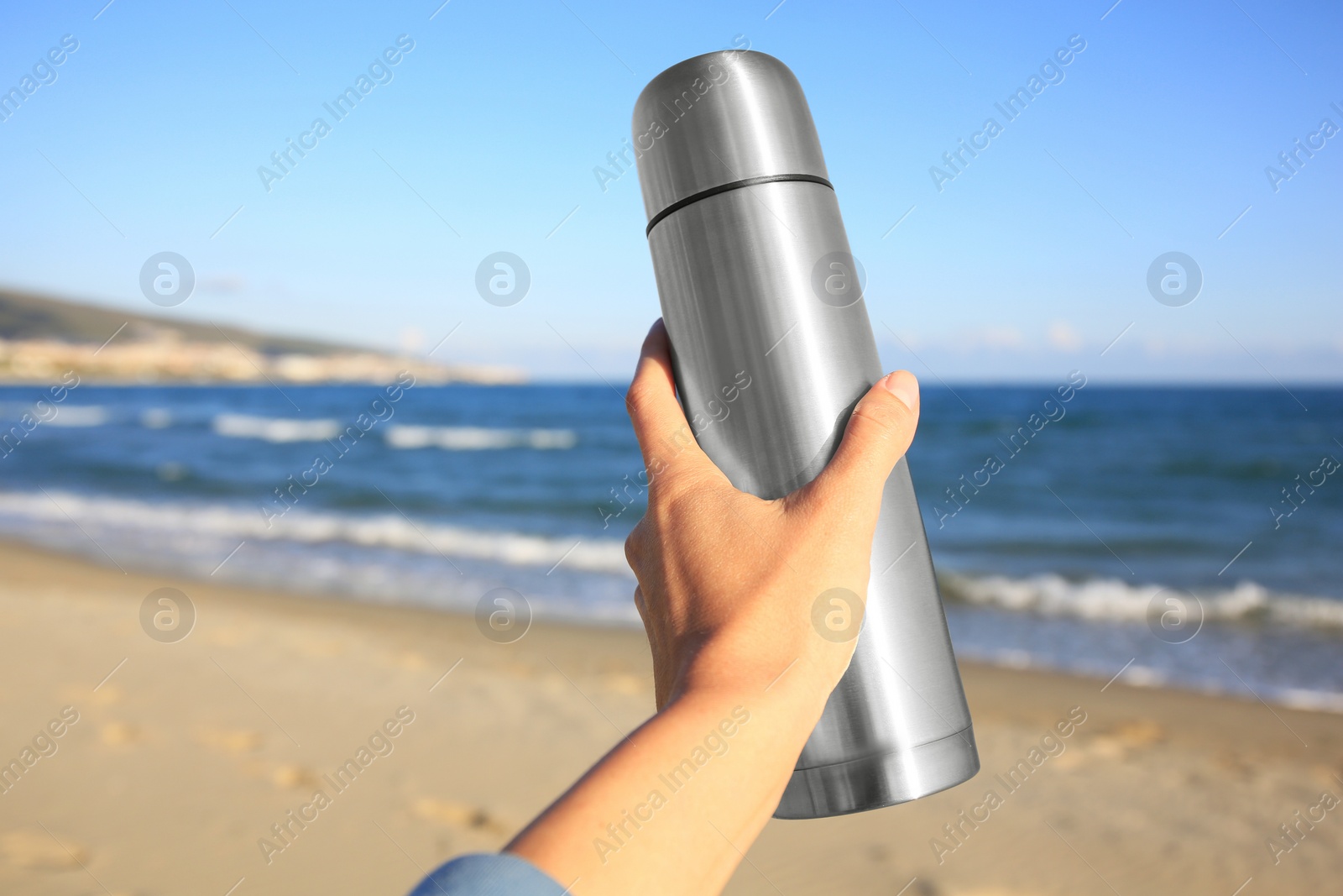 Photo of Woman holding metallic thermos with hot drink on beach near sea, closeup. Space for text