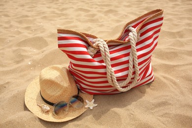 Stylish striped bag with straw hat, sunglasses, seashell and starfish on sand. Beach accessories