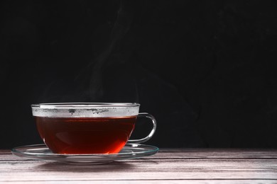 Glass cup of tea and saucer on wooden table against black background, space for text