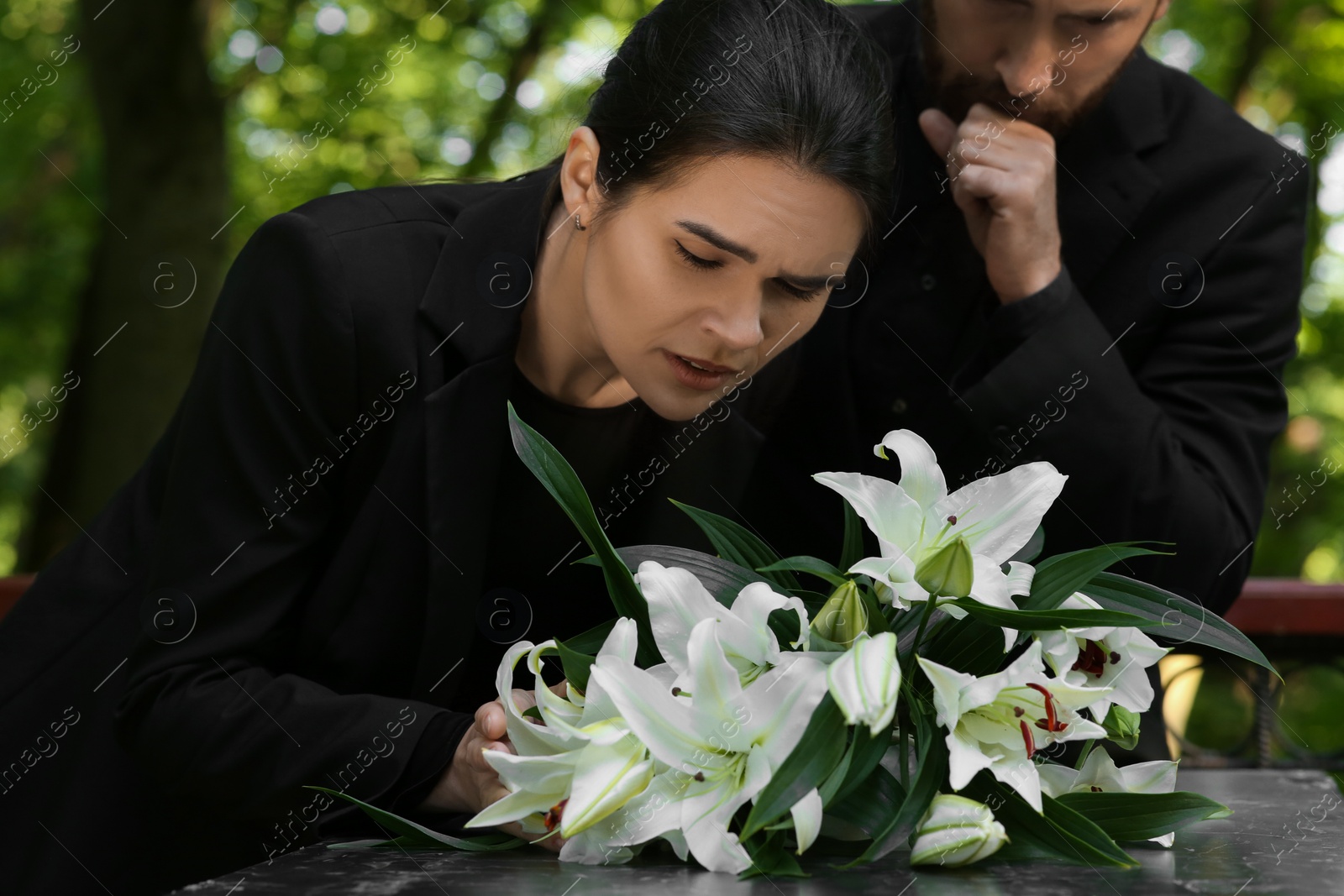 Photo of Sad couple mourning near granite tombstone with white lilies at cemetery. Funeral ceremony