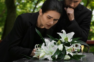 Sad couple mourning near granite tombstone with white lilies at cemetery. Funeral ceremony
