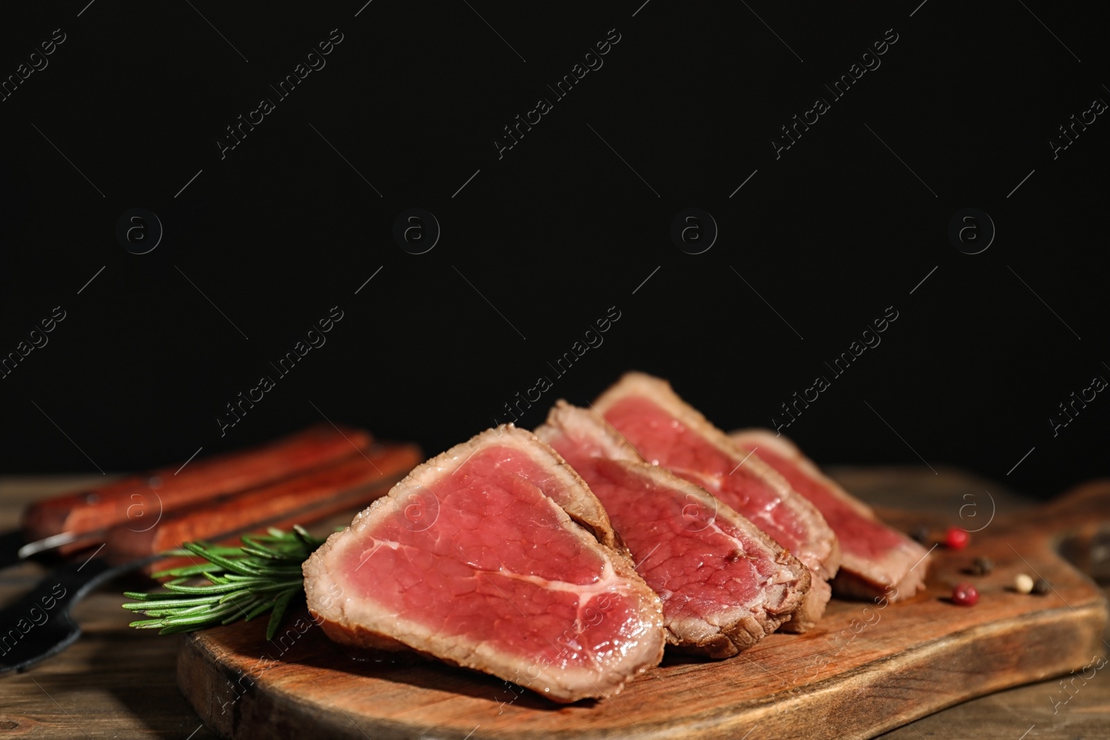 Photo of Board with roasted meat and rosemary on wooden table against black background. Space for text