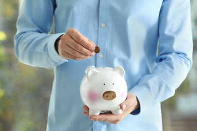 Man putting coin into piggy bank on blurred background, closeup. Money savings