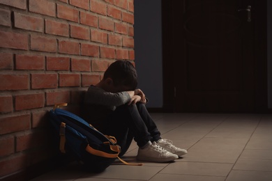 Photo of Sad little boy with backpack sitting on floor near brick wall indoors