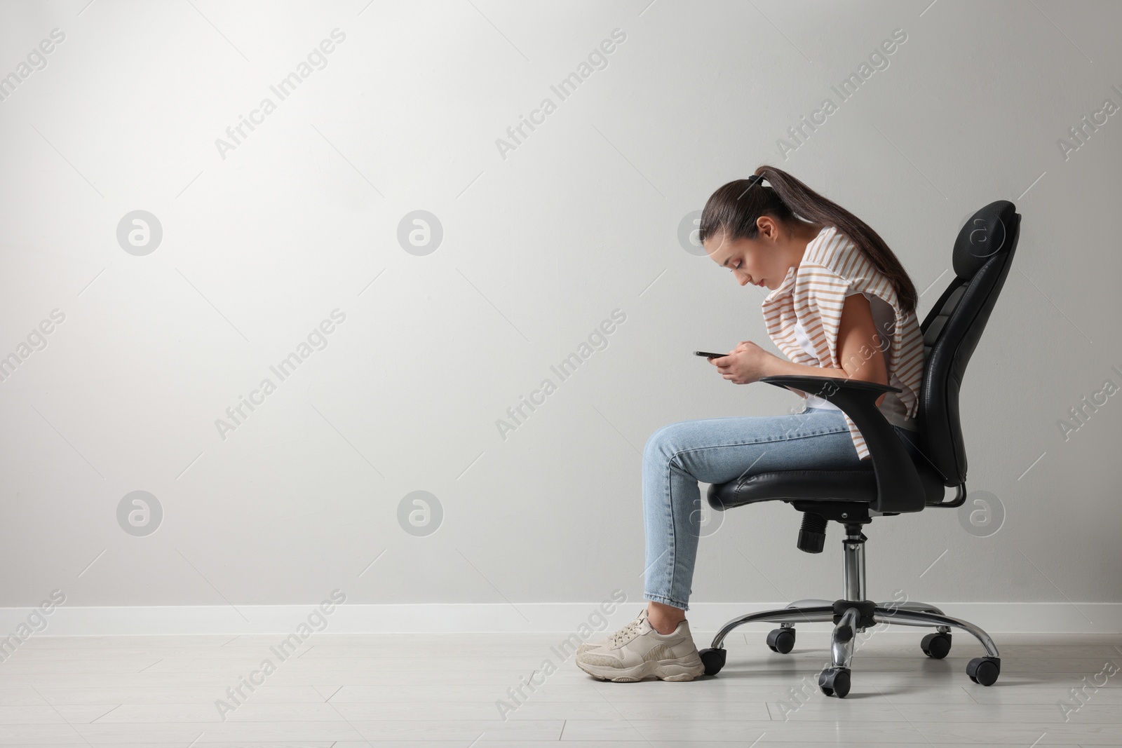 Photo of Young woman with poor posture using smartphone while sitting on chair near grey wall, space for text
