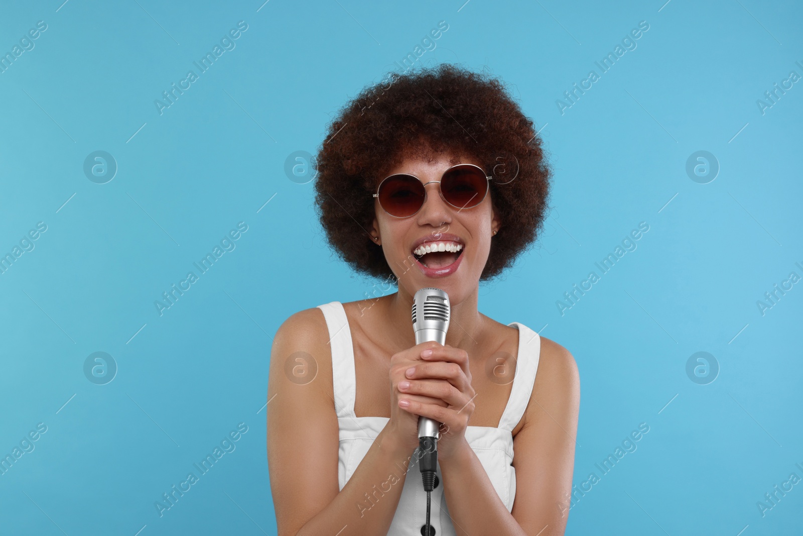 Photo of Curly young woman in sunglasses with microphone singing on light blue background