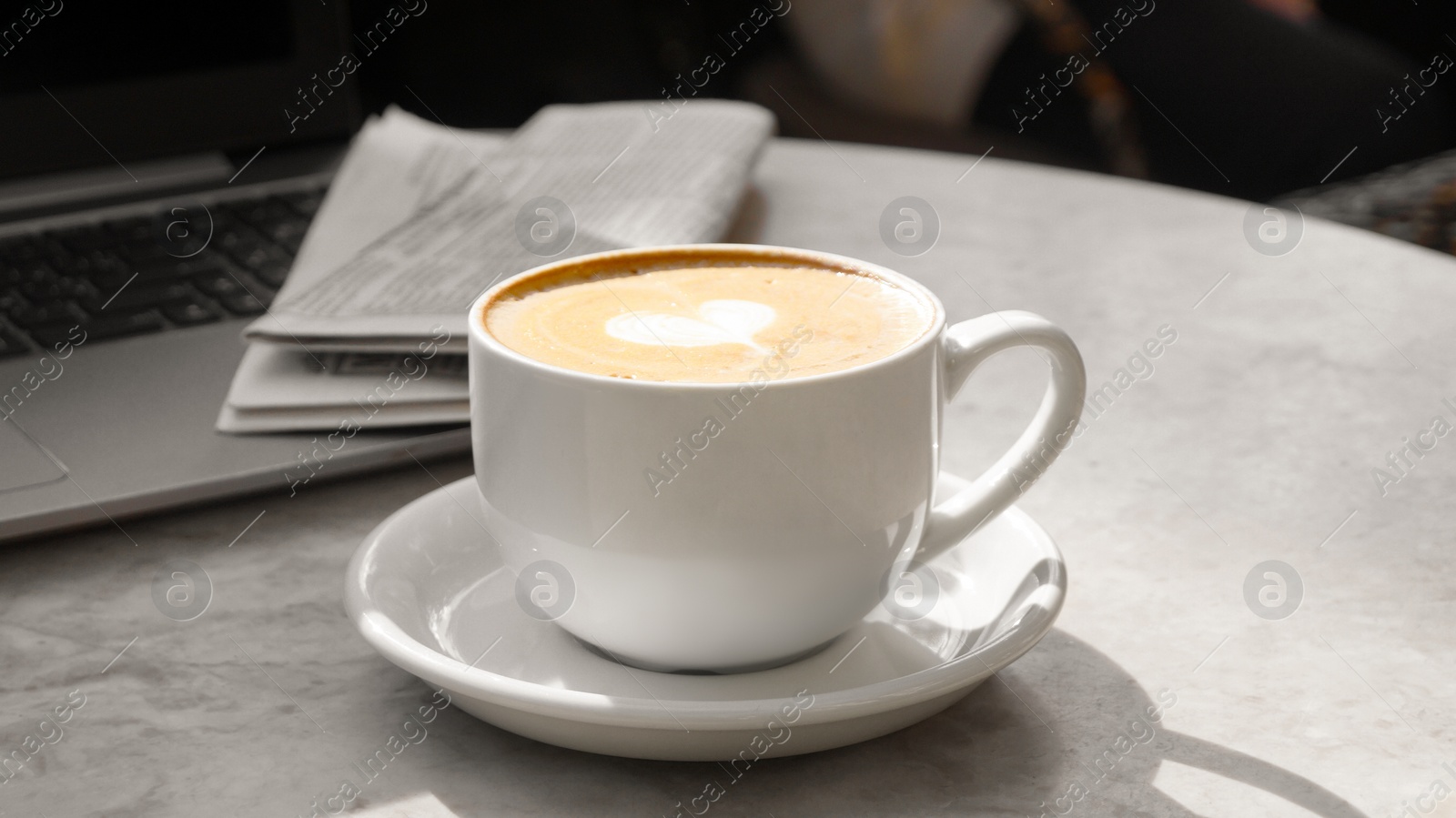 Photo of Cup of delicious coffee, newspaper and laptop on beige marble table outdoors