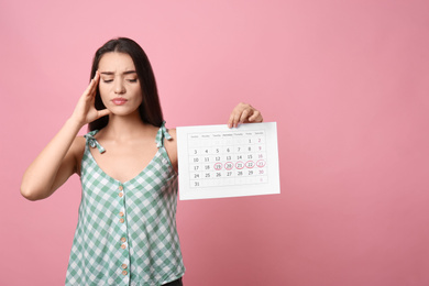 Young woman holding calendar with marked menstrual cycle days on pink background