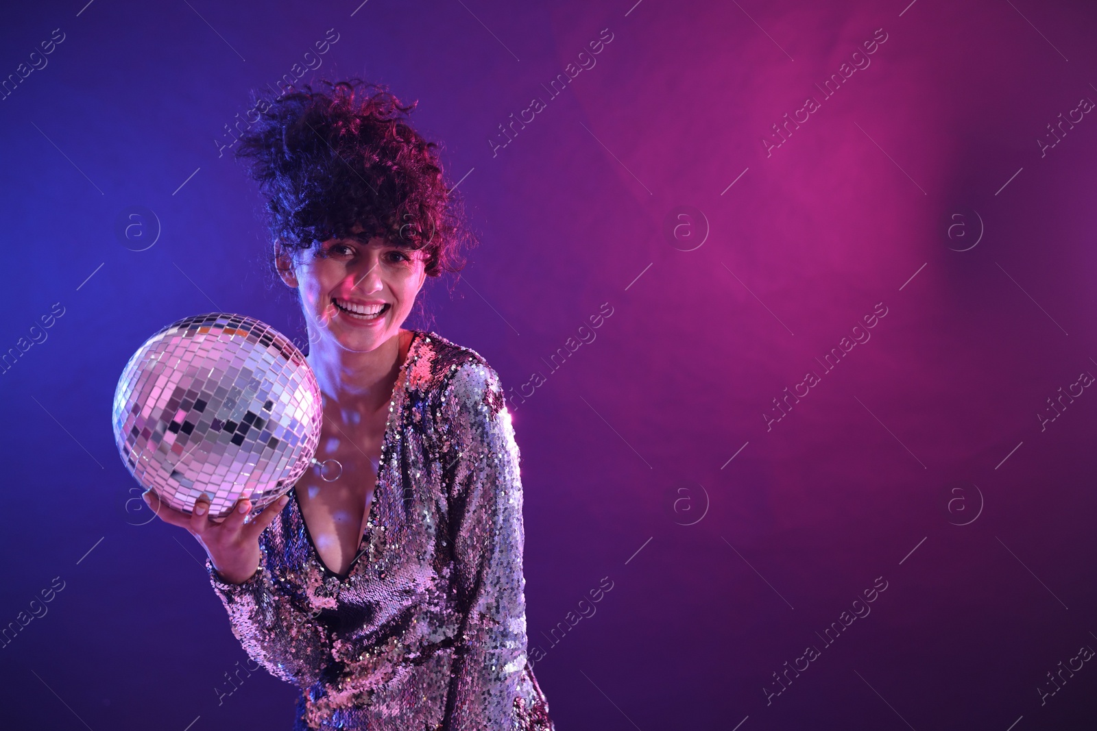 Photo of Beautiful young woman with disco ball posing on color background in neon lights. Space for text