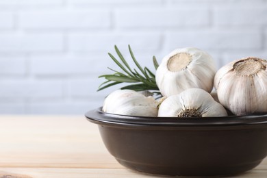 Photo of Fresh garlic bulbs in bowl and rosemary on wooden table, closeup. Space for text