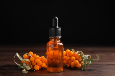 Photo of Ripe sea buckthorn and bottle of essential oil on wooden table against black background