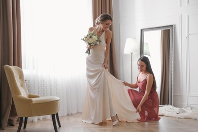 Gorgeous bride in beautiful wedding dress and her friend near mirror in room