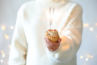 Photo of Woman holding cupcake with burning sparklers against blurred festive lights, closeup